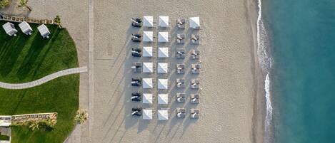 An aerial view of a beach with rows of sun loungers and umbrellas, a pathway, and a building with a pool.