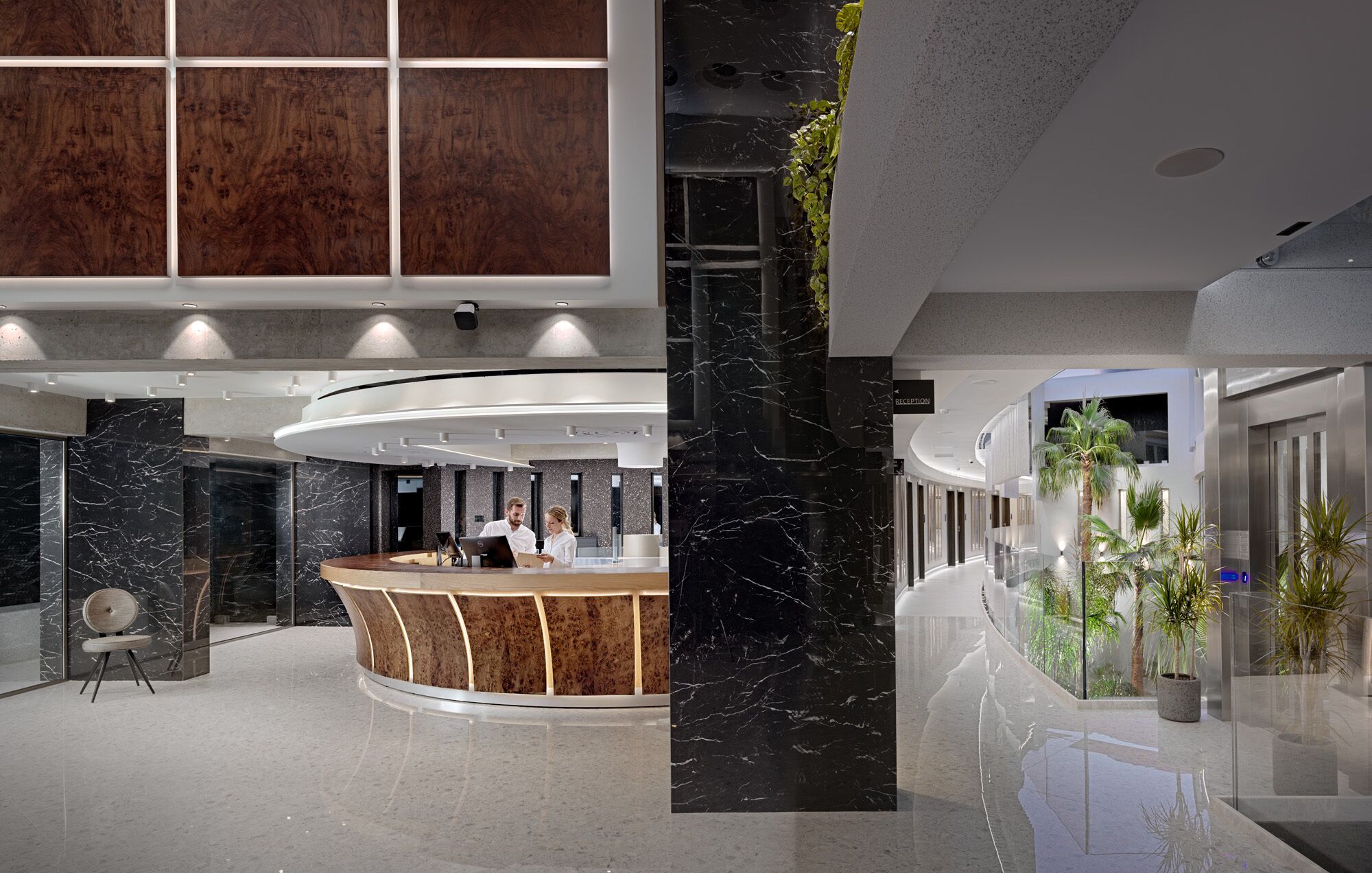 Un área de recepción moderna con un mostrador de madera curvo, un pilar de mármol negro y una pared de vidrio con plantas.