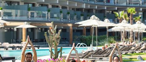 Tres personas haciendo yoga sobre césped frente a un hotel con piscina y palmeras.