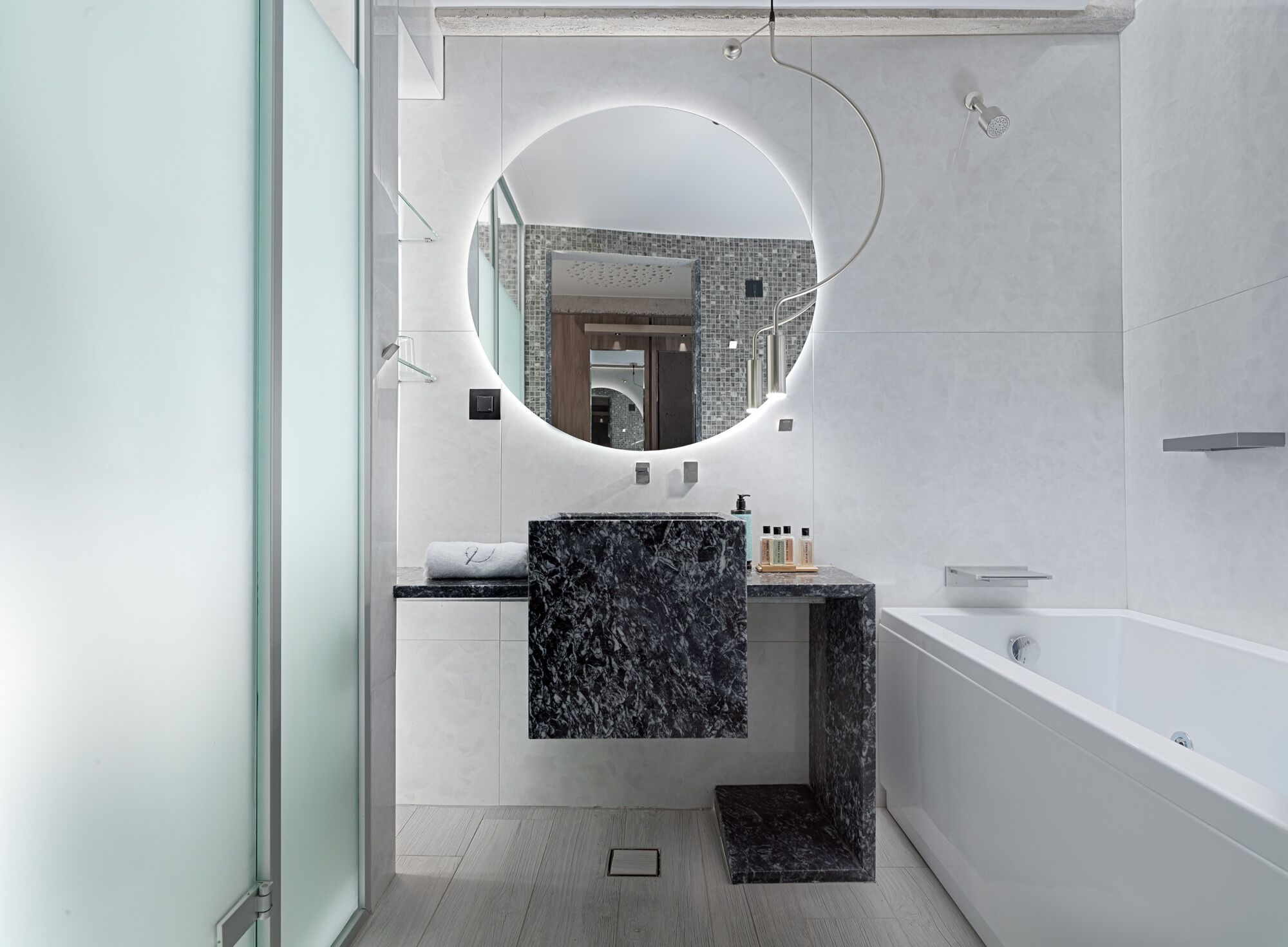 A modern bathroom with a round mirror, a marble sink, and a white bathtub.