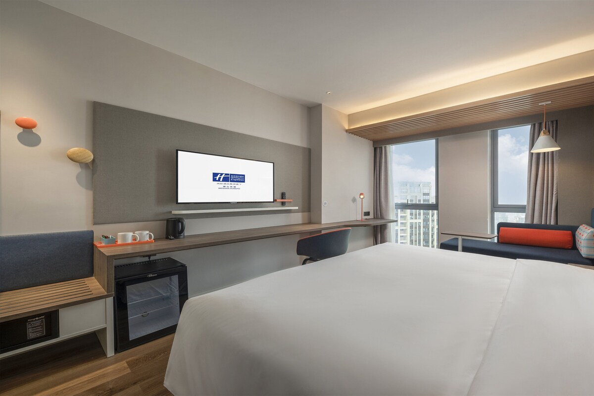 A modern hotel room with a large bed, a desk with a flat-screen TV, a seating area with a view of the city, and a small kitchenette.