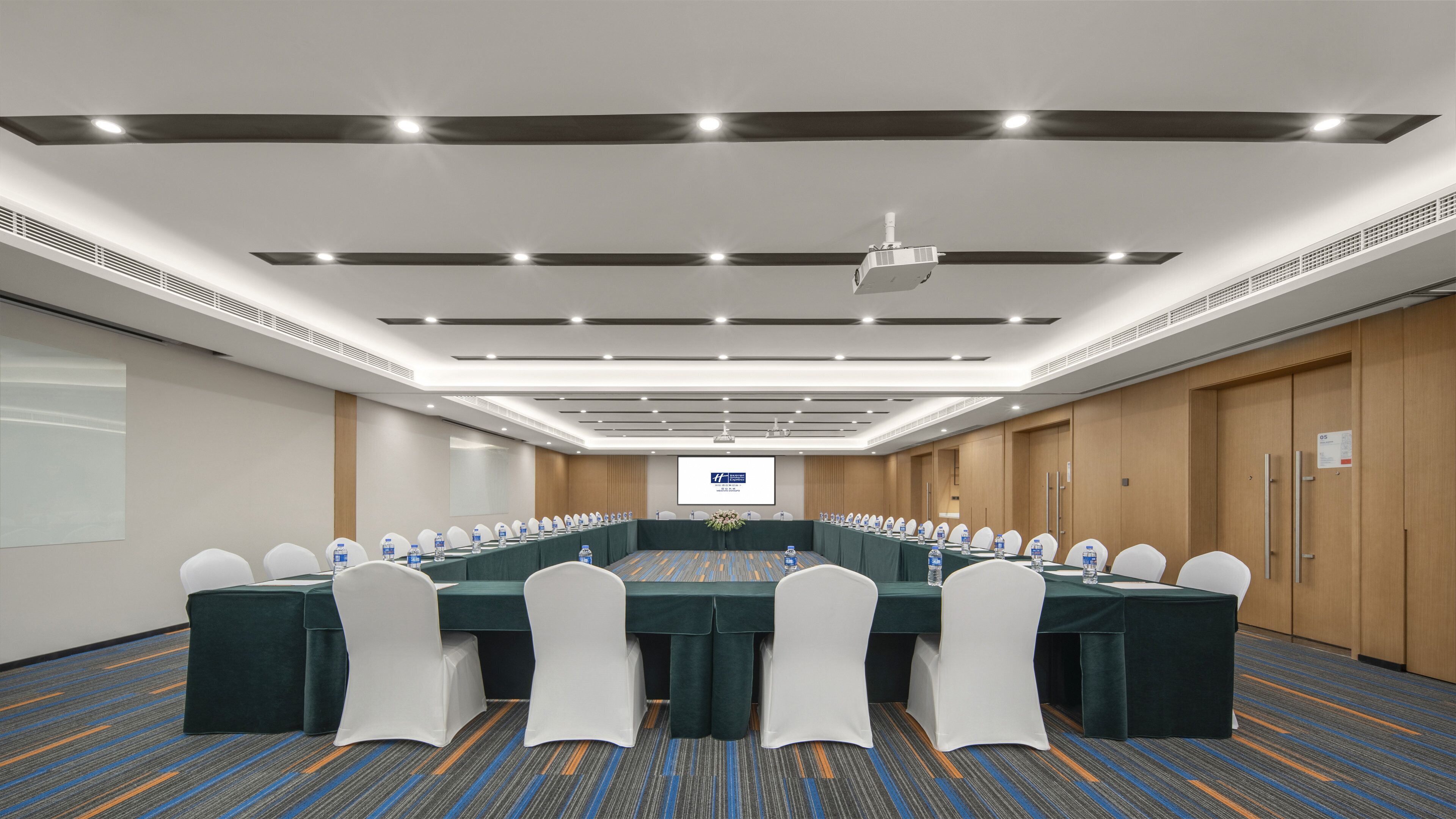 A conference room with a long table, white chairs, and a projector screen.