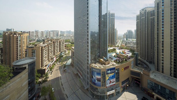 Un immeuble de grande hauteur doté d’une façade moderne en verre, entouré d’immeubles résidentiels de taille moyenne et d’une zone commerciale avec des enseignes bien visibles.