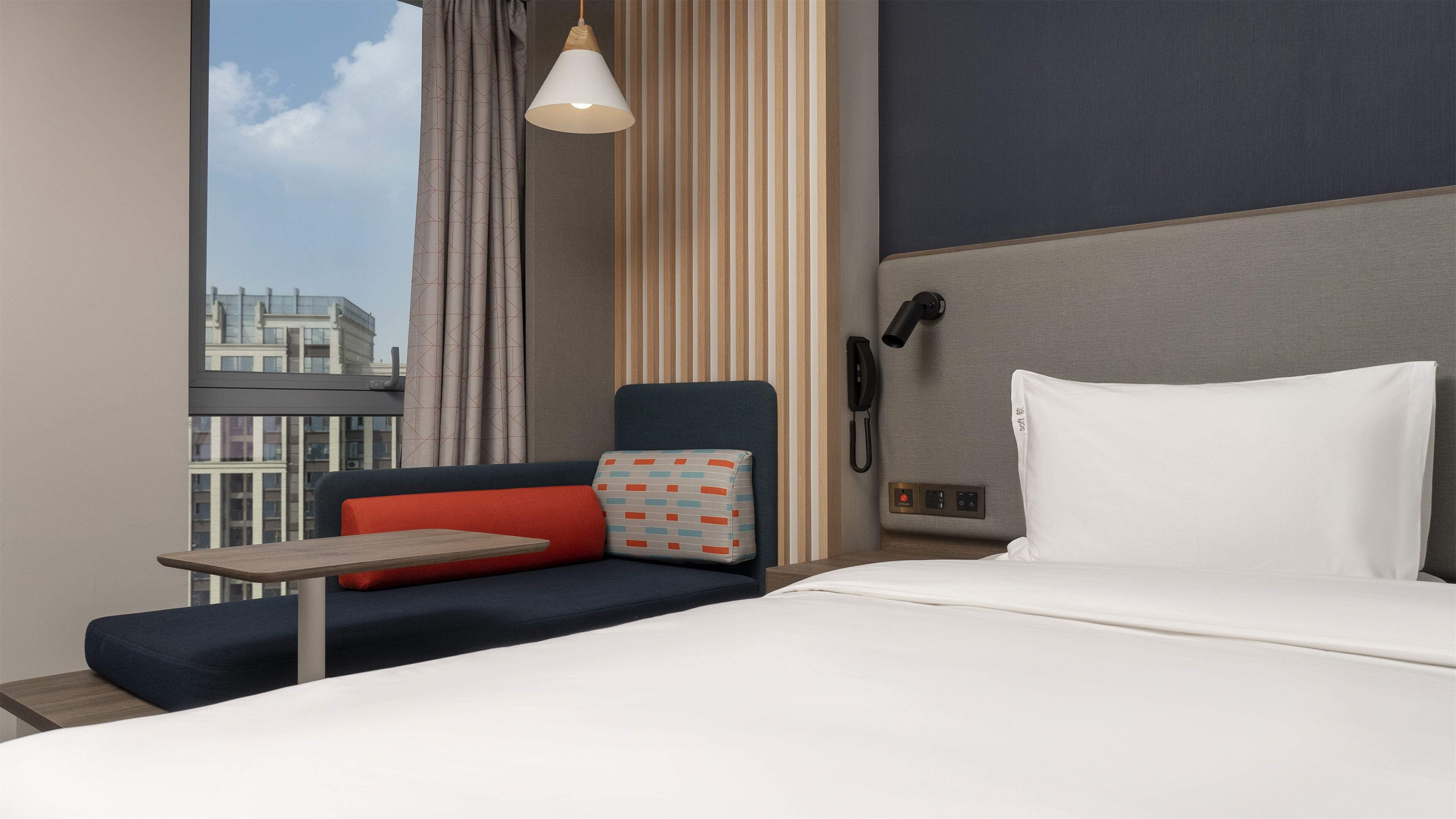 A hotel room with a bed, a desk, a chair, a pillow, a lamp, a phone, and a view of the city.