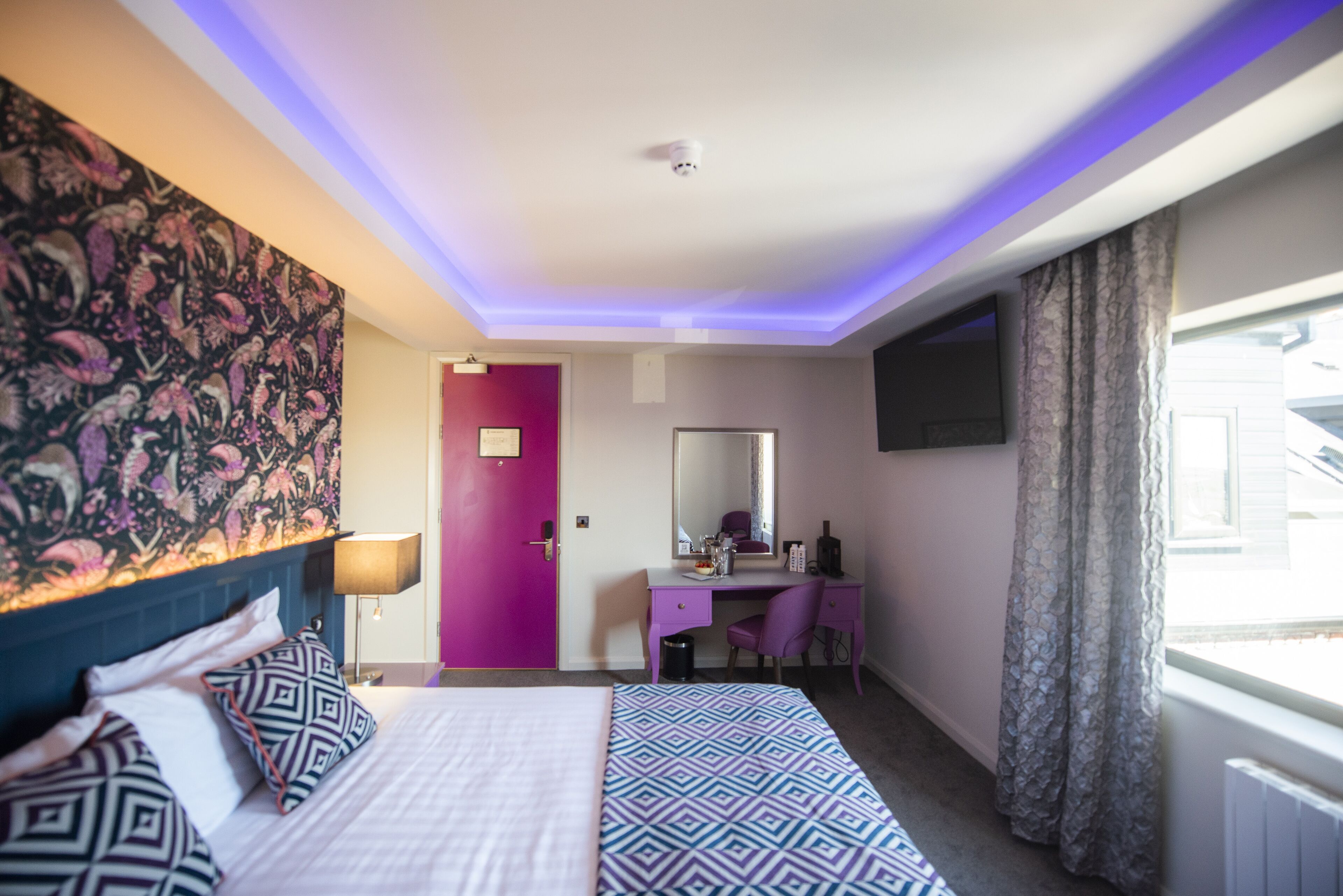A hotel room with a bed, a desk, a chair, a large mirror, and a window with curtains.