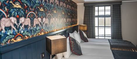 A hotel room with a large bed, a headboard with a patterned mural, a bedside table with a lamp, and a window with curtains.