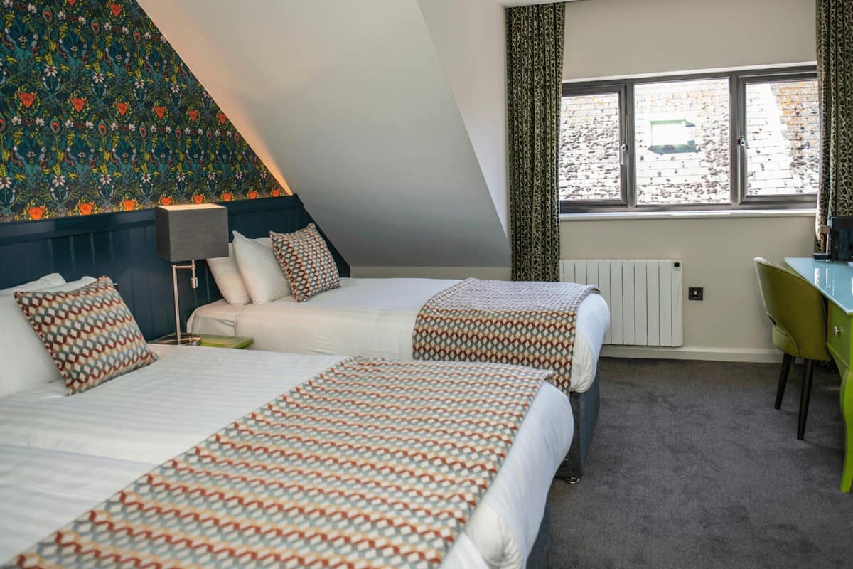 A hotel room with two beds, a desk, and a window with a view of buildings.