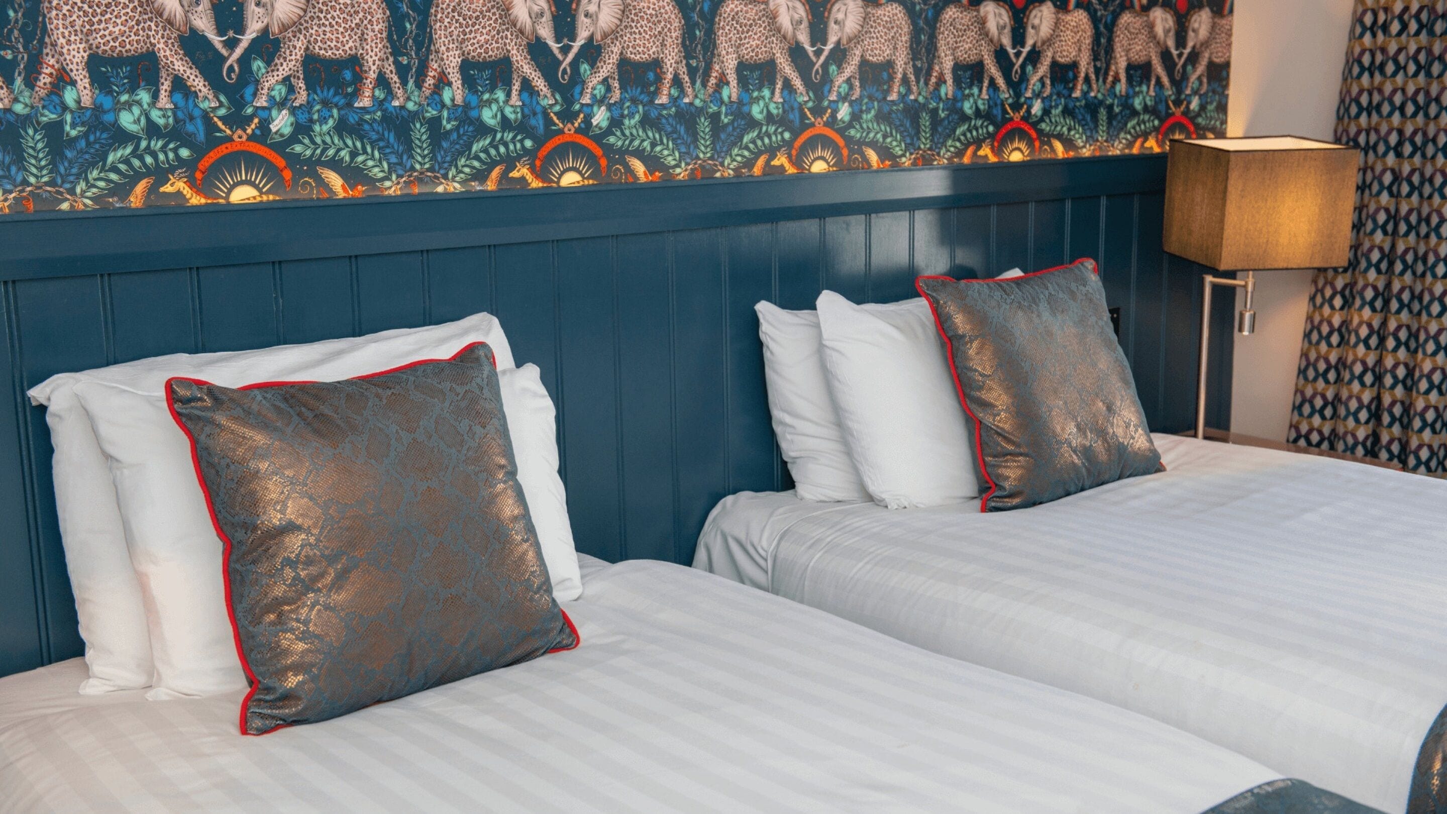 A hotel room with two beds, each with white linens and decorative pillows. The room features a blue headboard and a wall mural depicting animals and nature.