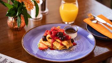 A plate of crepes topped with strawberries and blackberries, a small bowl of syrup, and a glass of orange juice on a wooden table.