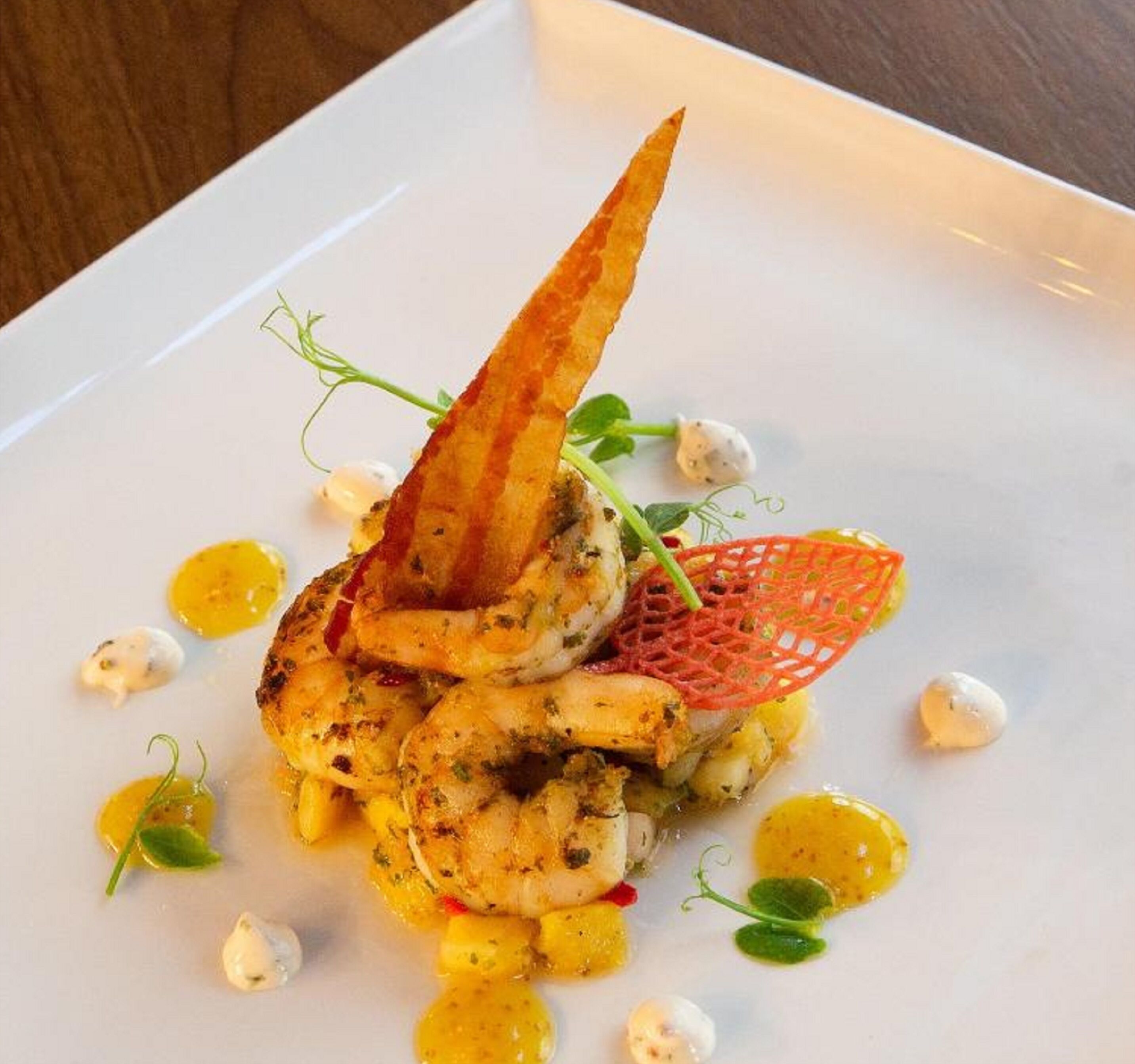 Un plato blanco con un guiso de mariscos, que incluye camarones y un trozo de pescado, adornado con hierbas y una estructura roja similar a una red.