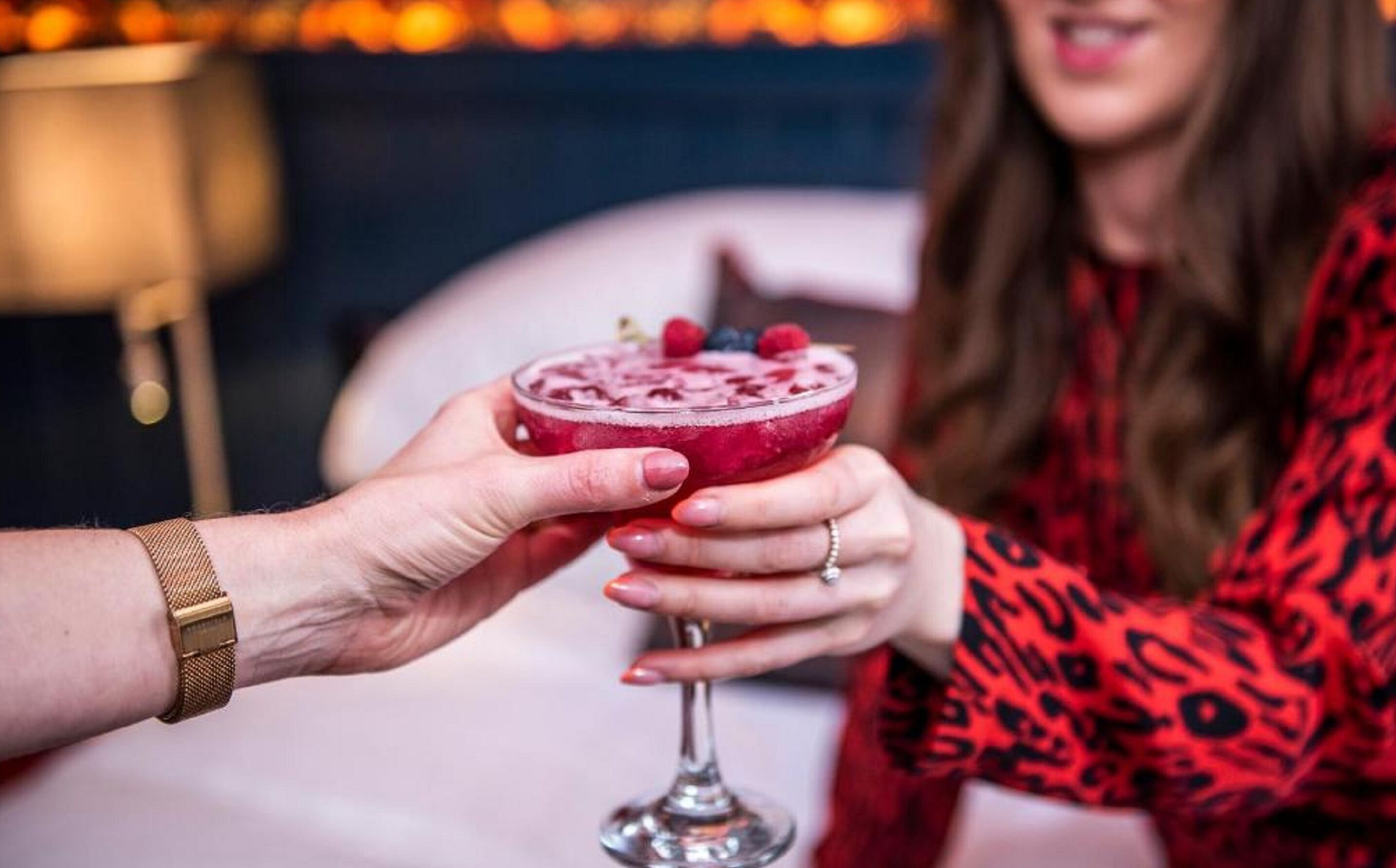 A person holding a cocktail glass with a fruit garnish, another person holding a similar glass, and a lamp in the background.