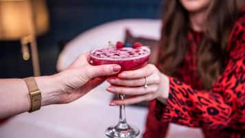 A person holding a cocktail glass with a fruit garnish, another person holding a similar glass, and a lamp in the background.