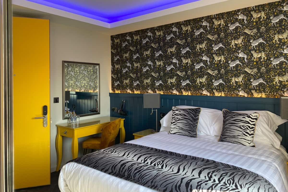A hotel room with a zebra-patterned bedspread, a desk with a chair, a mirror, and a wall with a patterned wallpaper.