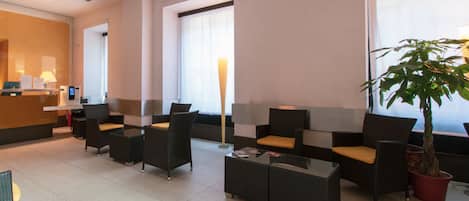 A hotel lobby with a reception desk, seating area with chairs and tables, a large potted plant, and large windows.