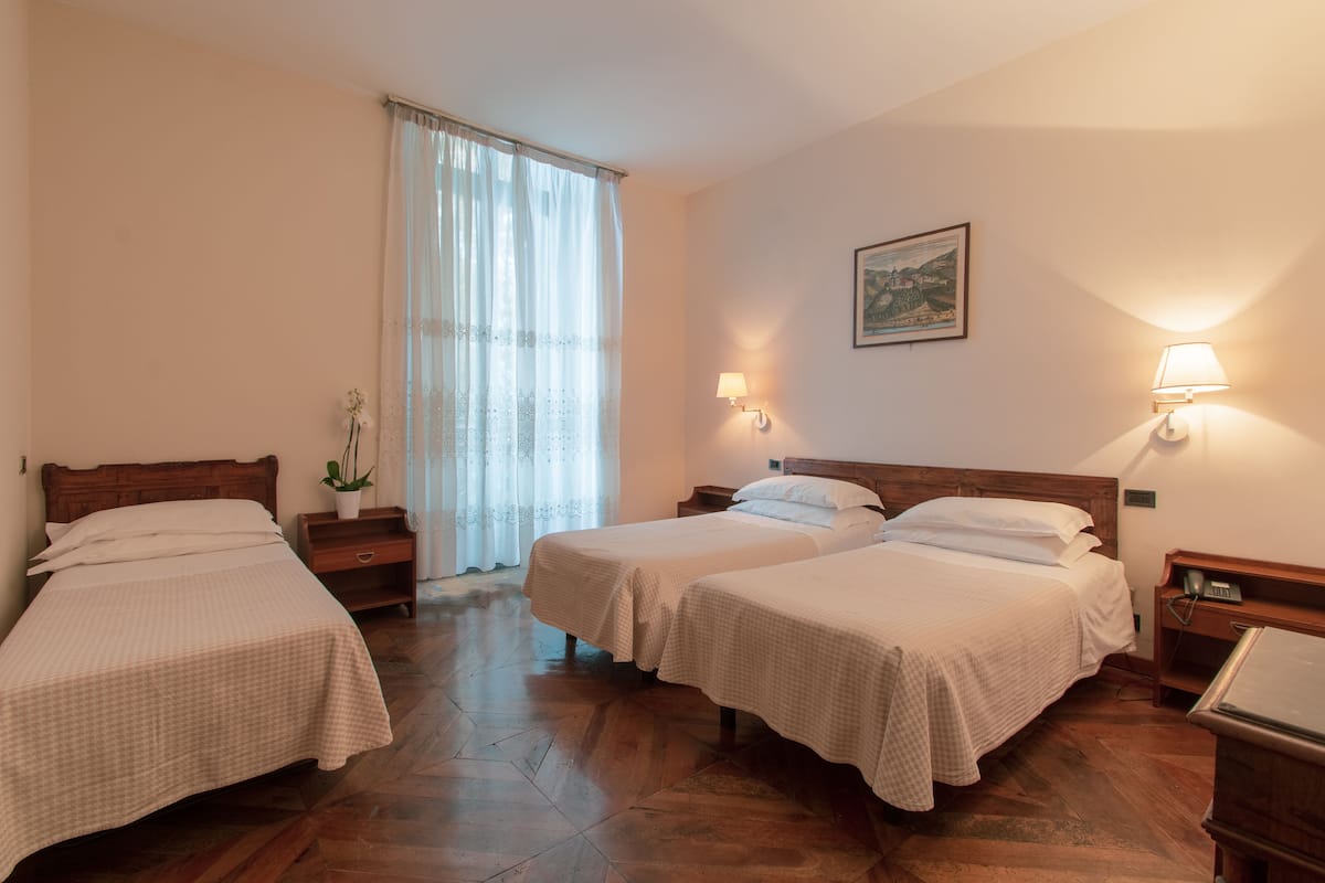 A hotel room with two single beds, a wooden floor, a window with sheer curtains, a bedside table with a lamp, and a painting on the wall.