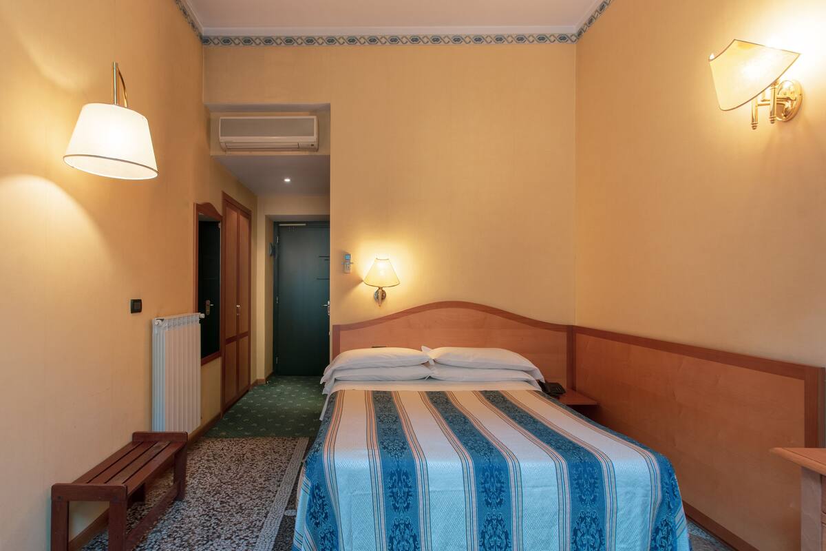 A hotel room with a bed, a bench, a radiator, and two wall-mounted lamps.