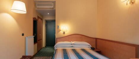 A hotel room with a bed, a bench, a radiator, and two wall-mounted lamps.