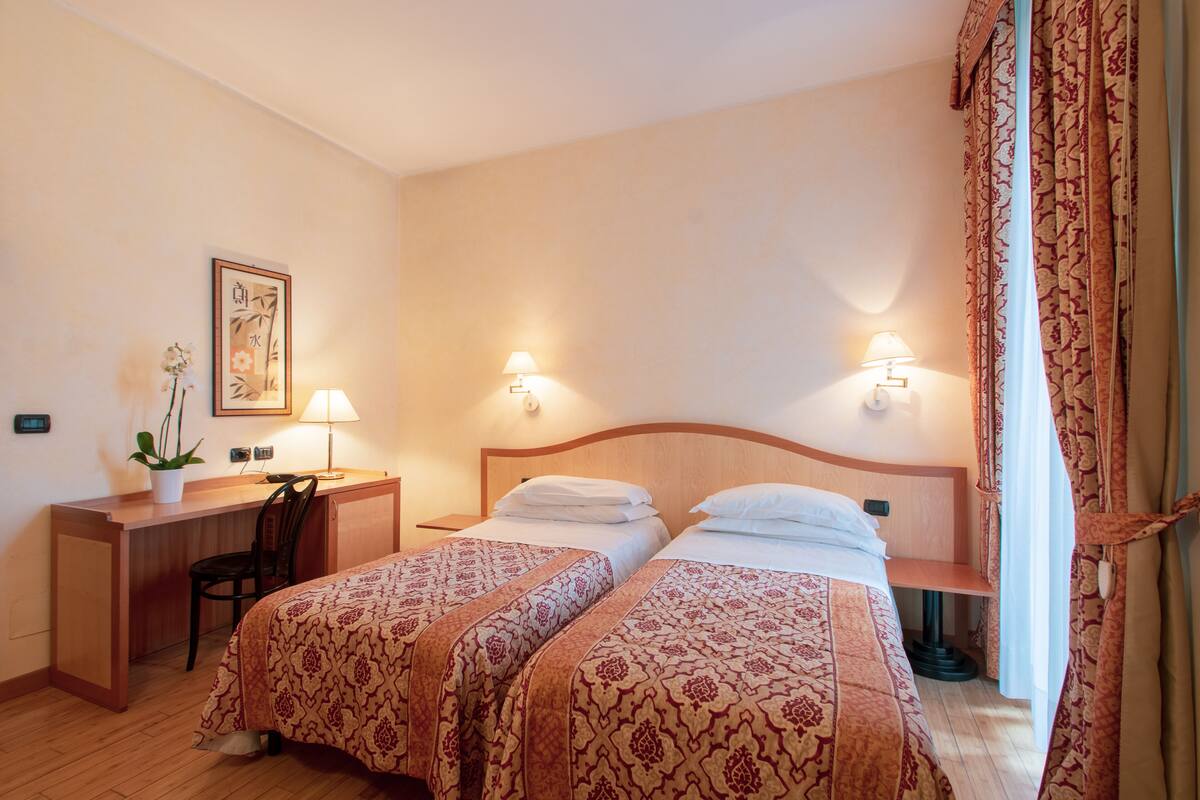 A hotel room with two beds, a desk, a chair, a lamp, and a framed picture on the wall.