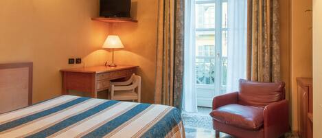 A hotel room with a bed, a desk with a lamp, a chair, a TV mounted on the wall, and a window with curtains.