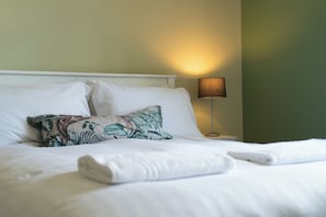 Una cama bien hecha con sábanas blancas y una almohada con estampado floral, una lámpara de noche y una mesita de noche blanca.