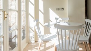 A dining area with a round table and white chairs, large windows, and a stone wall. - Toast Lettings | Crossgate, 2 bedroom Durham City cottage with castle views (Saint Margarets Garth)