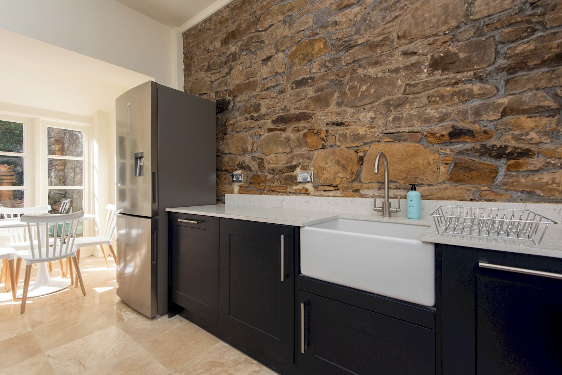 A kitchen with a stone wall backsplash, a stainless steel refrigerator, and a white sink with a soap dispenser.