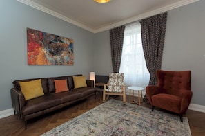 Una sala de estar con un sofá de cuero marrón, un sillón estampado, un cuadro abstracto y una alfombra con diseño.