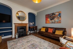 A living room with a fireplace, a brown leather sofa, a colorful abstract painting, and a large mirror on the wall. - Toast Lettings | Crossgate, 2 bedroom Durham City cottage with castle views (Saint Margarets Garth)