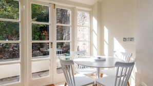A dining area with a round table and white chairs, large windows with a view of greenery, and a stone wall. - Toast Lettings | Crossgate, 2 bedroom Durham City cottage with castle views (Saint Margarets Garth)