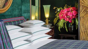 A neatly made bed with white pillows and a plaid bedspread, a green upholstered headboard, a bedside table with a lamp, and a vase of pink flowers.