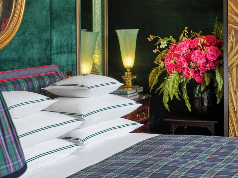 A neatly made bed with white pillows and a plaid bedspread, a green upholstered headboard, a bedside table with a lamp, and a vase of pink flowers.