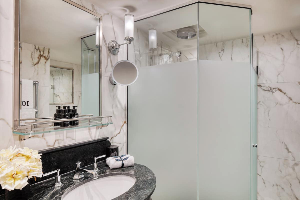 A modern bathroom with a glass shower enclosure, a marble-tiled wall, a glass shelf with toiletries, and a granite sink.