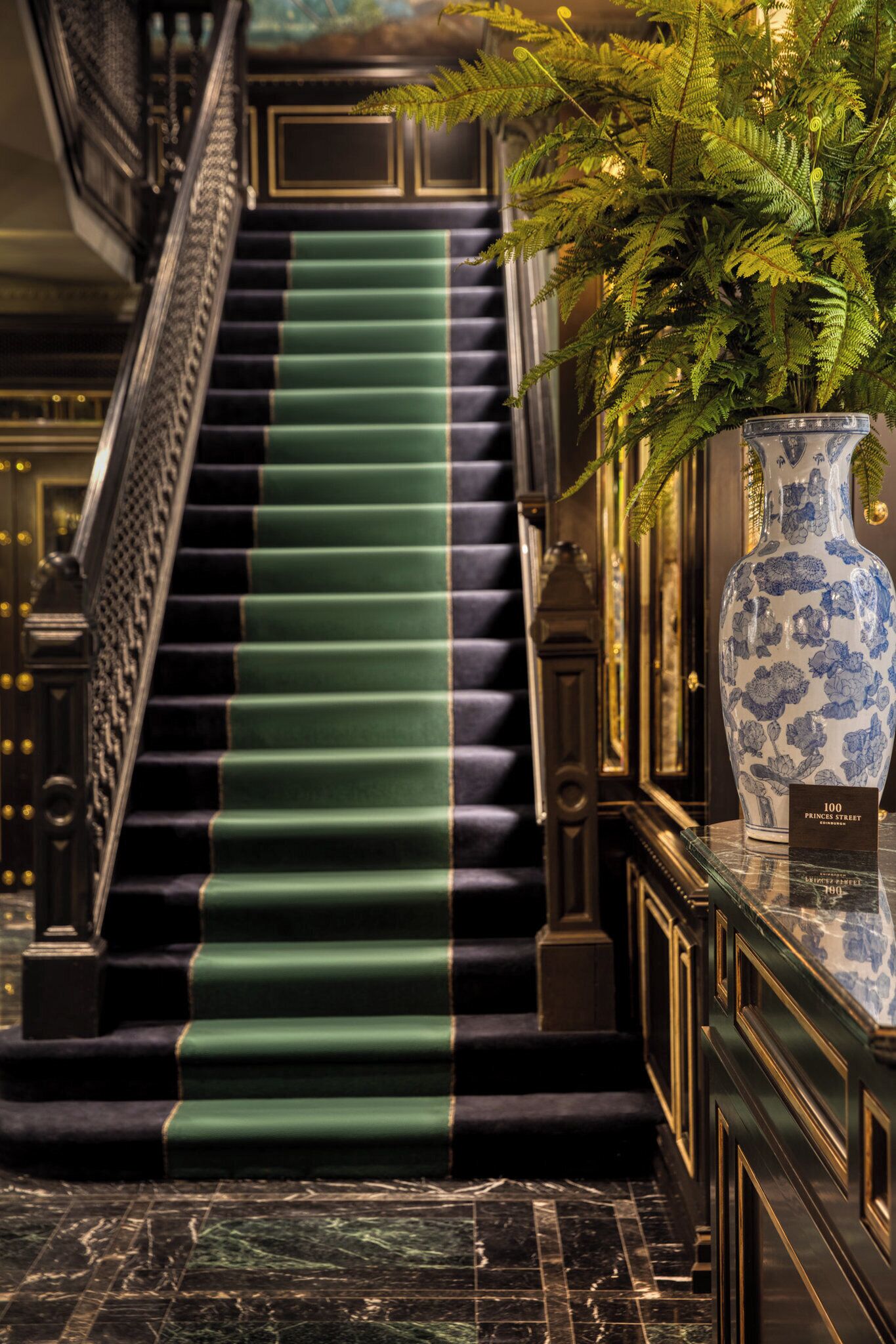 Una imponente escalera con peldaños cubiertos de moqueta verde, un gran jarrón azul y blanco, y un letrero que indica '100 Prince Street'.
