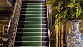 Una imponente escalera con peldaños cubiertos de moqueta verde, un gran jarrón azul y blanco, y un letrero que indica '100 Prince Street'.
