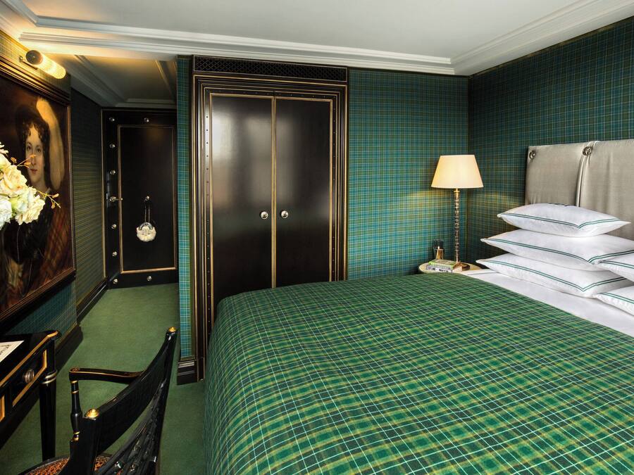 Ein Hotelzimmer mit einem großen Bett, einem Schreibtisch mit Blumenvase, einem Sessel, einem Kleiderschrank und einem Nachttisch mit Lampe.