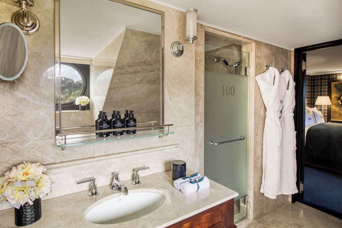 A modern bathroom with a large mirror, a glass shelf with toiletries, a sink, and a shower area.