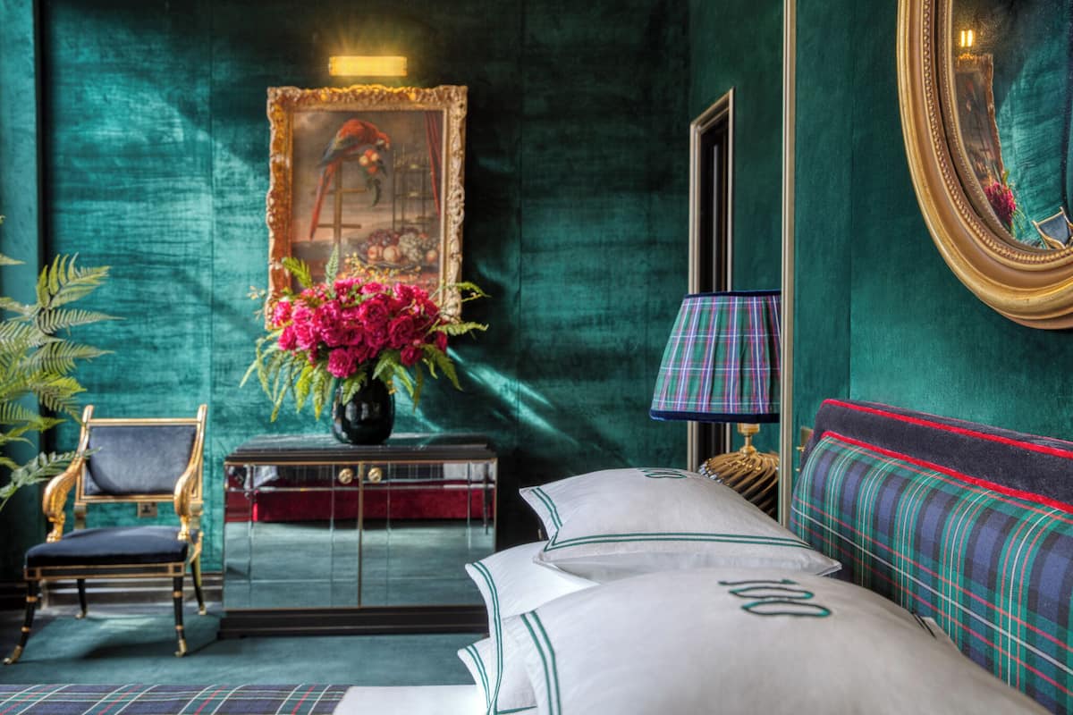 A room with a bed, a chair, a table with a vase of flowers, a mirror, and a painting on the wall.