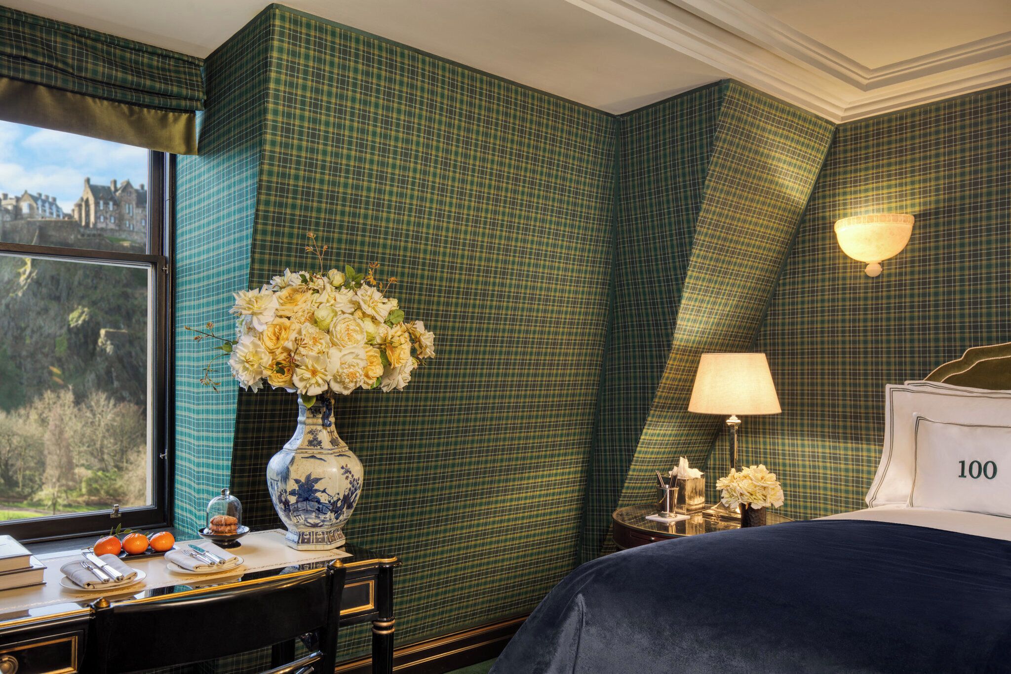 Une chambre d’hôtel avec un mur vert à carreaux, une grande fenêtre donnant sur un beau paysage, un lit avec le chiffre « 100 » inscrit sur la tête de lit, un vase rempli de fleurs et un bureau avec une lampe.