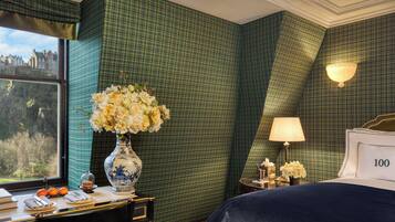 Une chambre d’hôtel avec un mur vert à carreaux, une grande fenêtre donnant sur un beau paysage, un lit avec le chiffre « 100 » inscrit sur la tête de lit, un vase rempli de fleurs et un bureau avec une lampe.