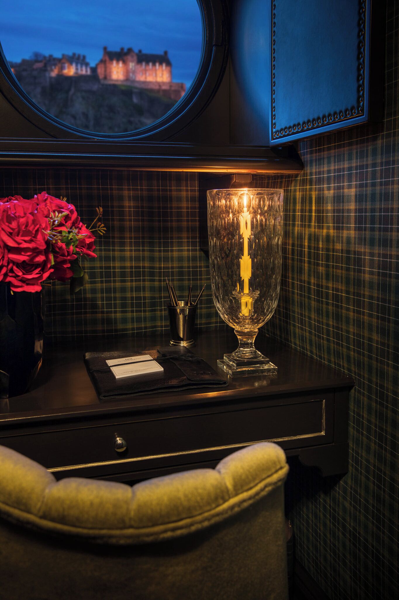 Ein Zimmer mit Blick auf ein Schloss, ein Fenster, ein Glaslaterne, eine Vase mit Blumen und eine Wand mit Karomuster.