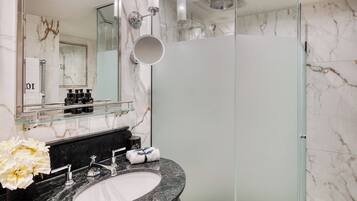 Un baño moderno con cabina de ducha de vidrio, pared revestida de mármol, repisa de vidrio con artículos de aseo y lavamanos de granito.
