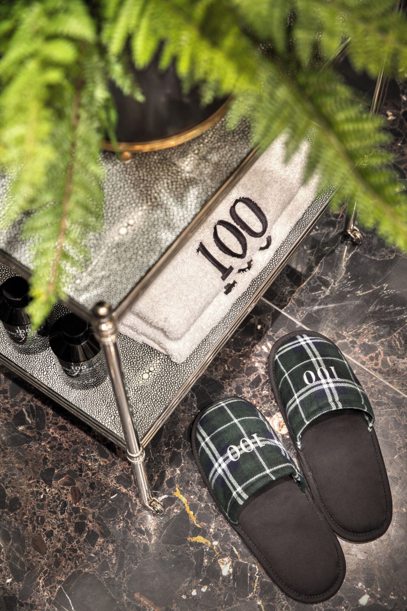 Un par de pantuflas escocesas sobre un suelo de mármol, una mesita con un adorno y una planta de hojas verdes.