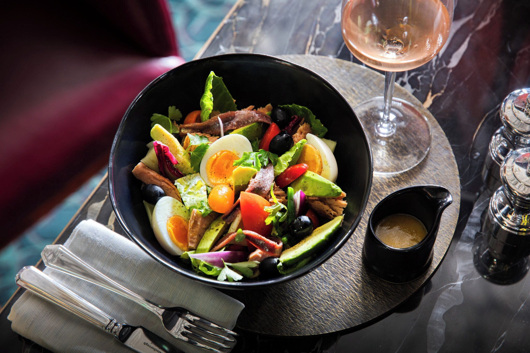A bowl of salad with a small cup of dressing on a slate plate, accompanied by a glass of wine and salt and pepper shakers.