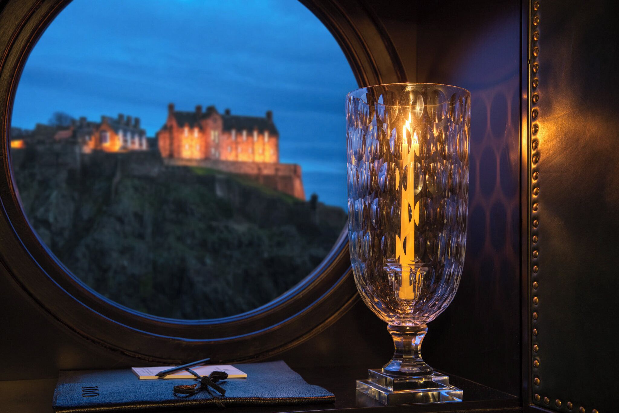 A view of a castle through a window, a glass table with a candle, and a leather-bound book.