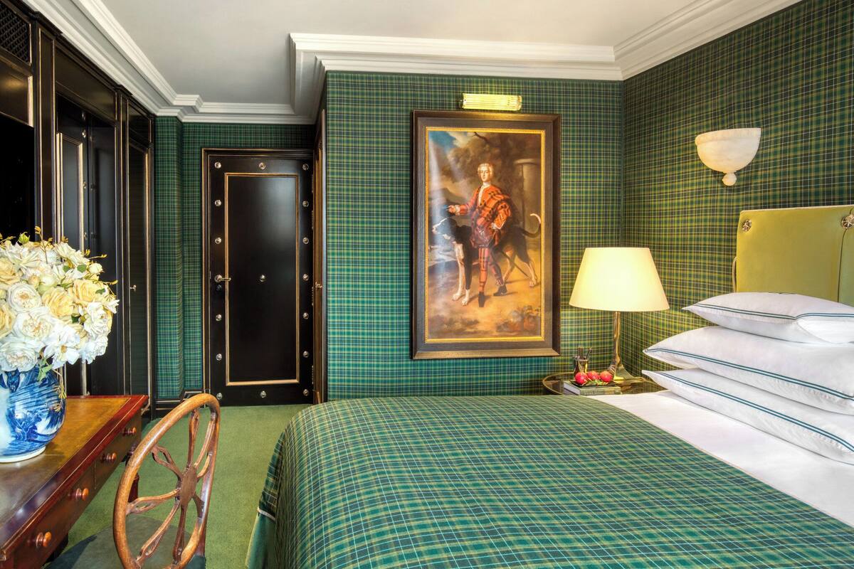 A hotel room with a green plaid wallpaper, a bed with white linens, a wooden chair, a vase with flowers, a painting on the wall, and a nightstand with a lamp.