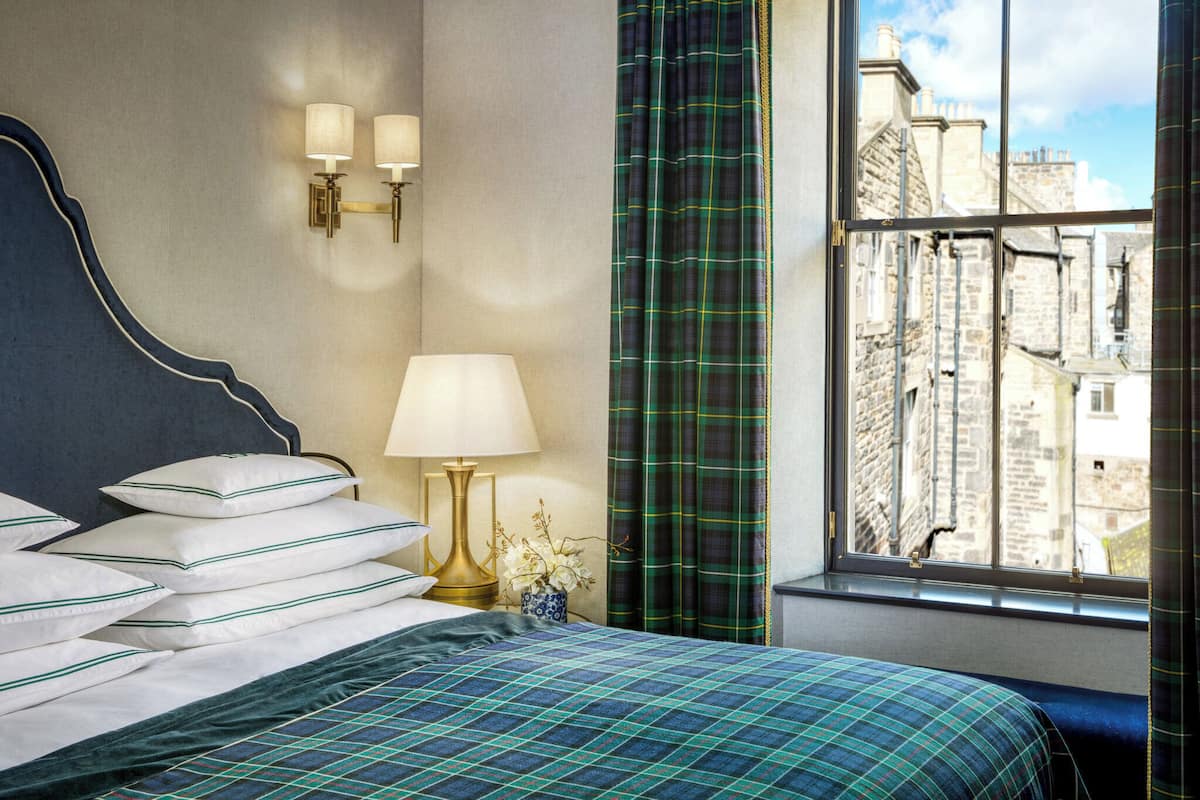 A neatly made bed with white pillows and a blue and green checkered bedspread, a gold bedside lamp, and a window with a view of buildings.