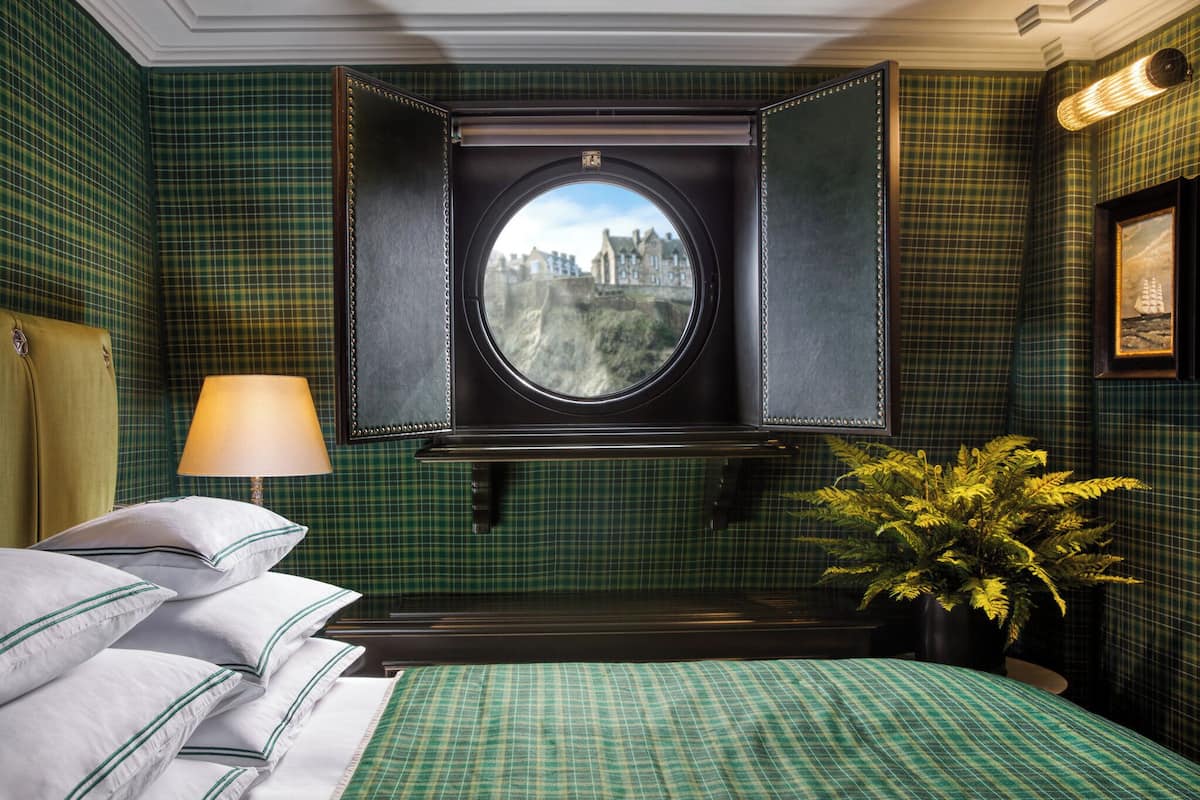 A room with a bed, pillows, a lamp, a potted plant, and a window with a view of a castle.