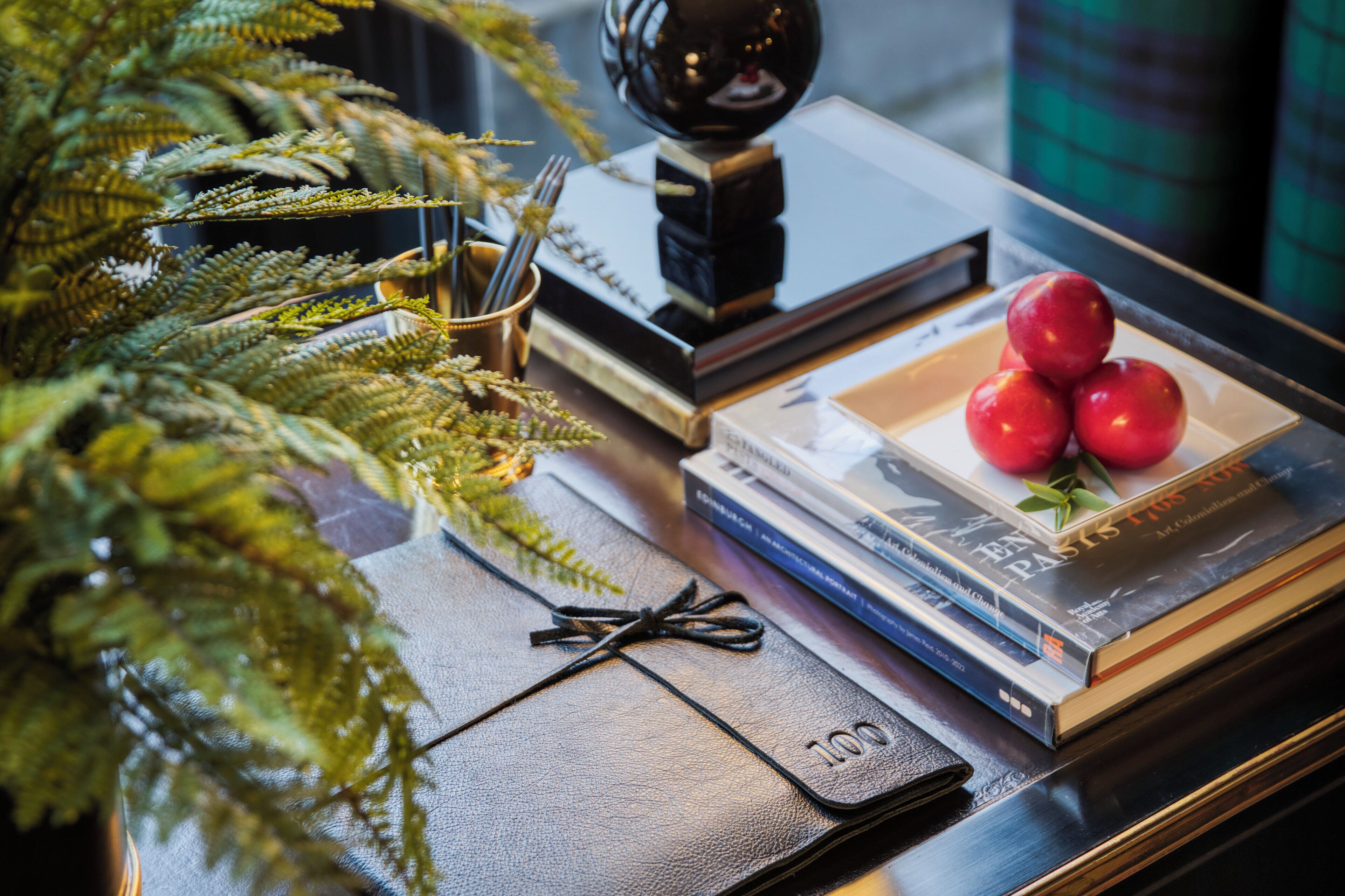 A table with a plant, books, a black leather notebook, and a plate with apples.