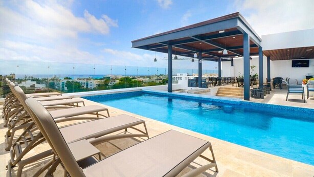 A rooftop pool with lounge chairs and a covered seating area, overlooking a cityscape. - 1BR Ocean View, Gym & Rooftop Bar! (Playa del Carmen)