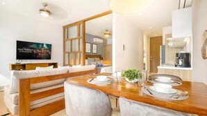 A modern dining area with a wooden table set for two, a sofa, and a TV mounted on the wall. - 1BR Ocean View, Gym & Rooftop Bar! (Playa del Carmen)